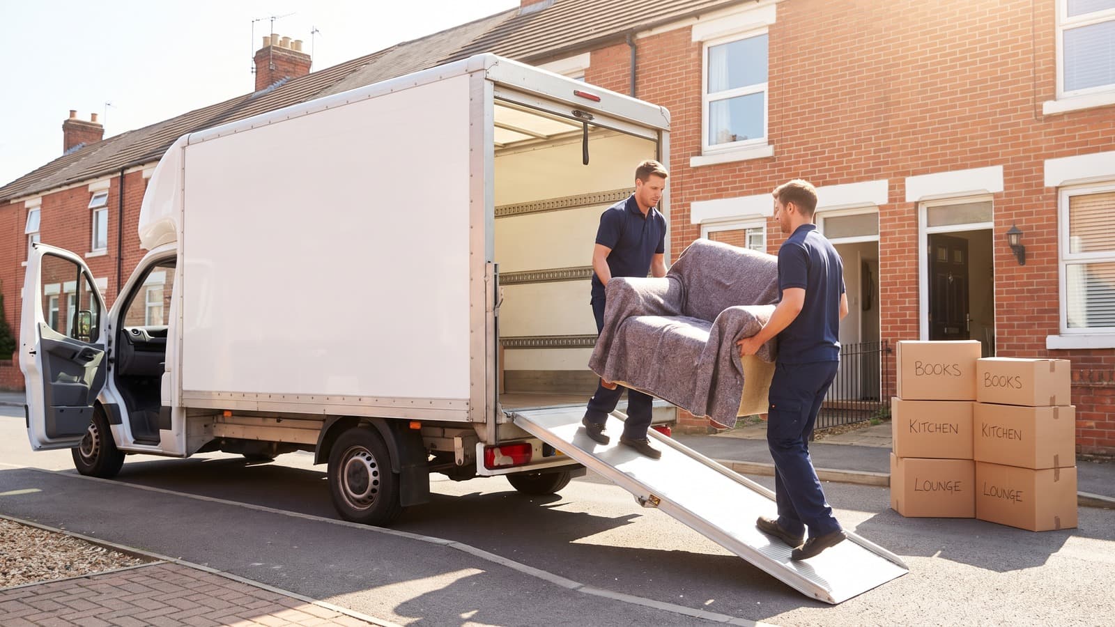 A Luton van reversed up to the open door of a terraced house early in the morning, crew carrying a wrapped armchair out at pace with labelled boxes stacked on the path