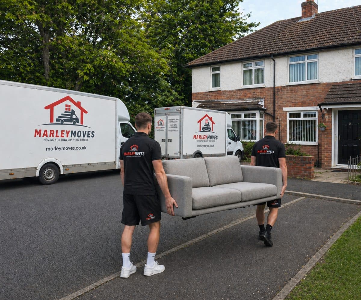 Two Marley Moves crew members in branded polos carrying a blanket-wrapped sofa with two Marley Moves branded vans behind, a typical domestic removal in Gillingham, Dorset